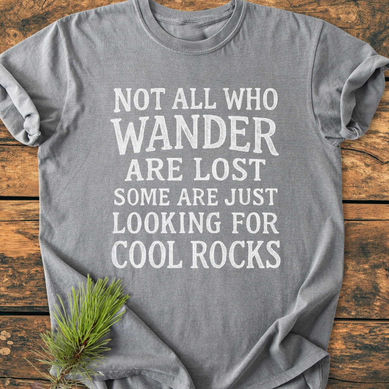 Gray Not All... Rustic Chalkboard - Vintage Wash Tee laid flat, featuring a large white distressed text: “NOT ALL WHO WANDER ARE LOST SOME ARE JUST LOOKING FOR COOL ROCKS” on a wooden surface.