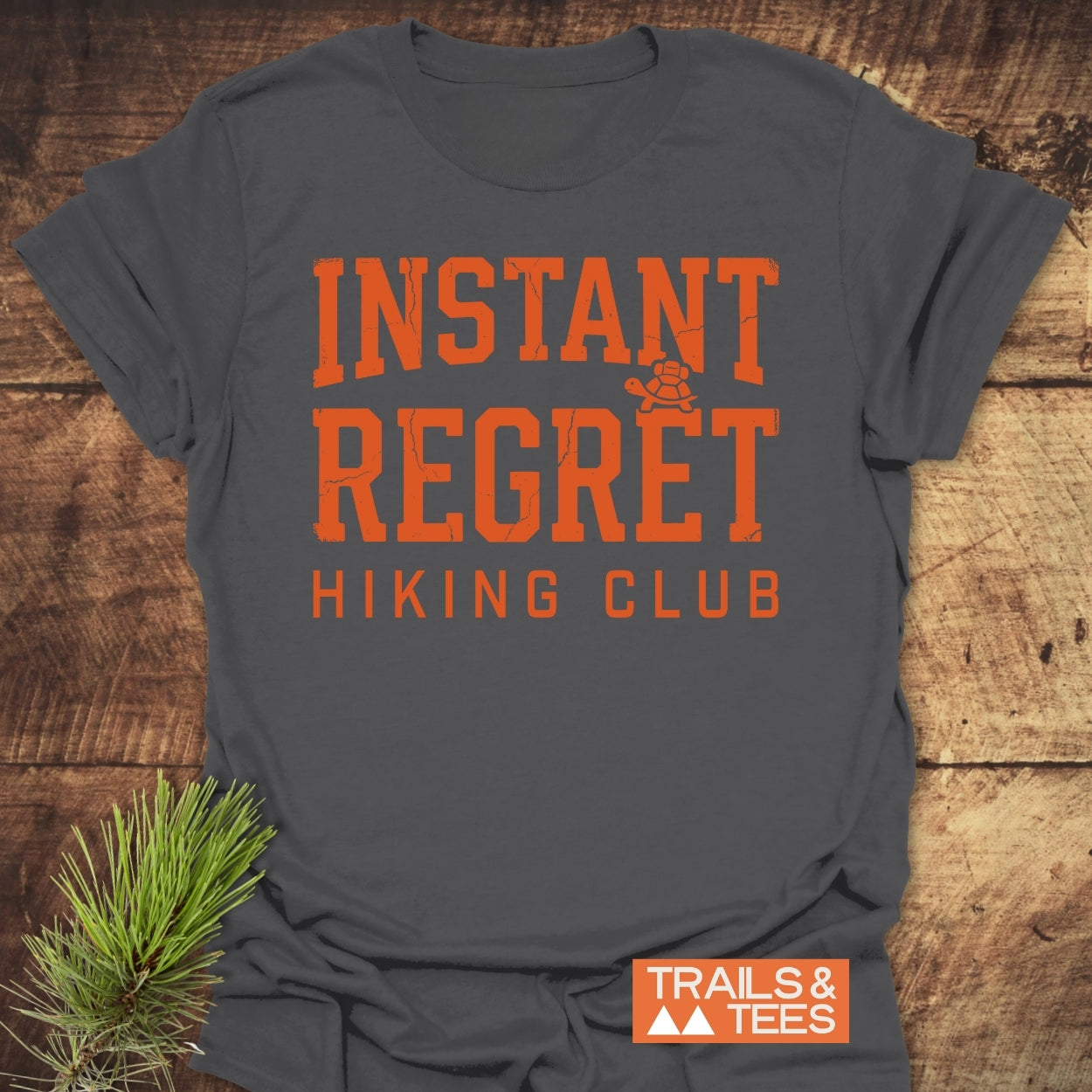 Instant Regret Hiking Club T-Shirt displayed on a wooden surface, features bold distressed orange lettering and a turtle icon. Includes the Trails & Tees logo, emphasizing adventure-ready comfort and design.