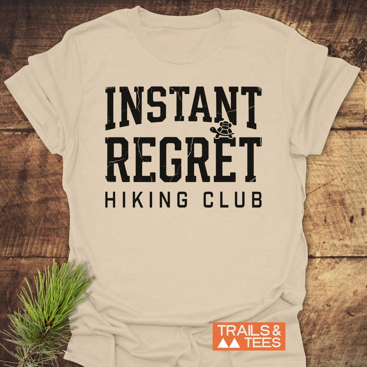 Instant Regret Hiking Club T-Shirt featuring distressed block letters and a turtle icon on soft, ring-spun cotton, laid on wood with pine needles. Includes orange TRAILS & TEES logo nearby.