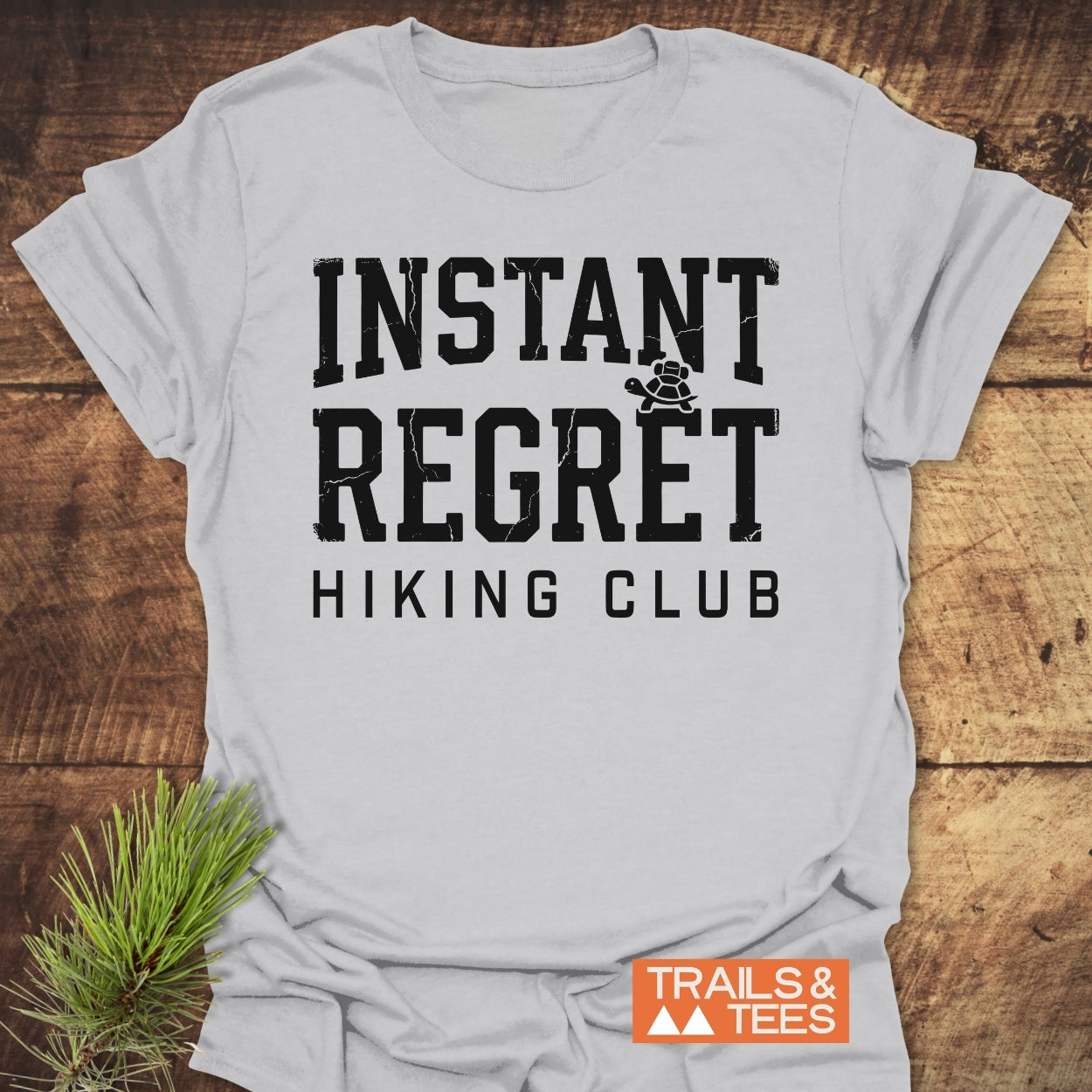 The Instant Regret Hiking Club T-Shirt features distressed black lettering and a turtle icon on light-gray fabric, laid flat on a tabletop, with an orange TRAILS & TEES logo nearby.