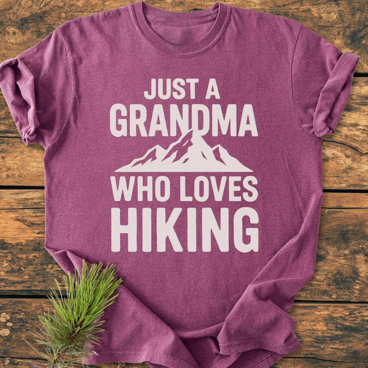 Heather-purple Grandma Loves Hiking - Vintage Wash Tee on wood surface, features bold white lettering and mountain graphic. Pine sprig accents the cozy, relaxed fit, 100% ring-spun cotton t-shirt.