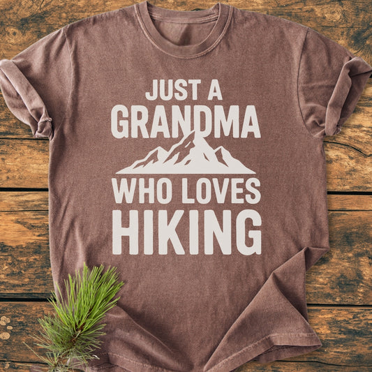 Grandma Loves Hiking - Vintage Wash Tee displayed on rustic wood, featuring bold white text and a mountain graphic. Accented by a pine sprig, showcasing its cozy, relaxed fit and durable, garment-dyed cotton fabric.