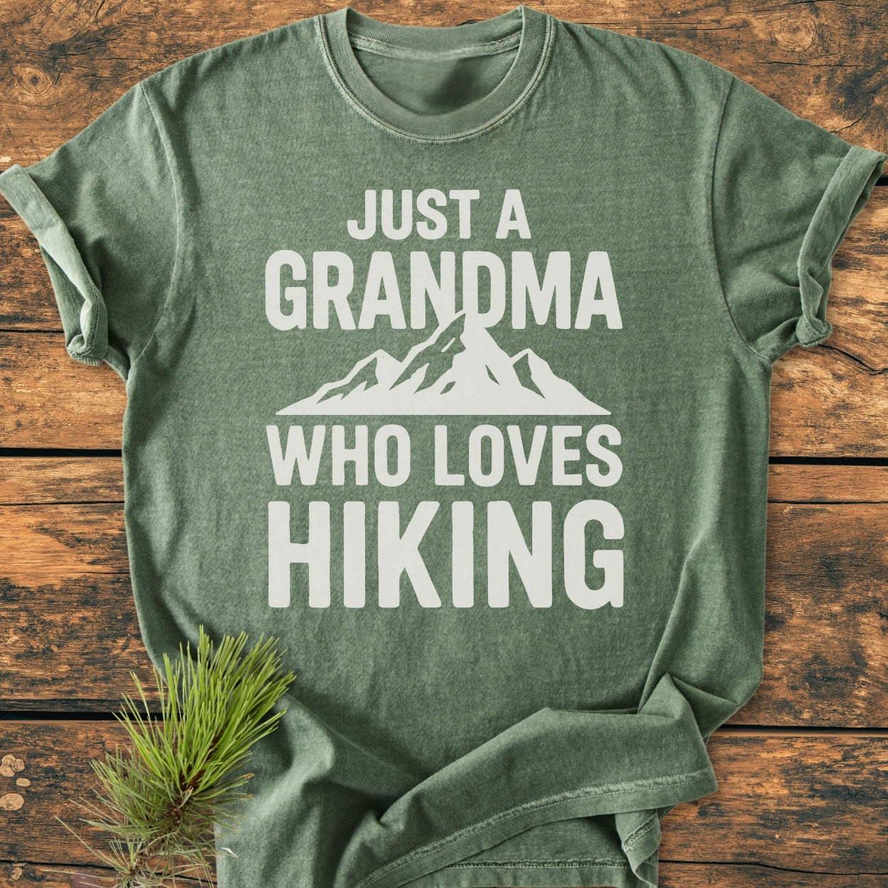 Grandma Loves Hiking - Vintage Wash Tee, green with “JUST A GRANDMA WHO LOVES HIKING” in white, features a mountain silhouette. Laid flat on wood with pine sprig, showcasing its relaxed, durable design.