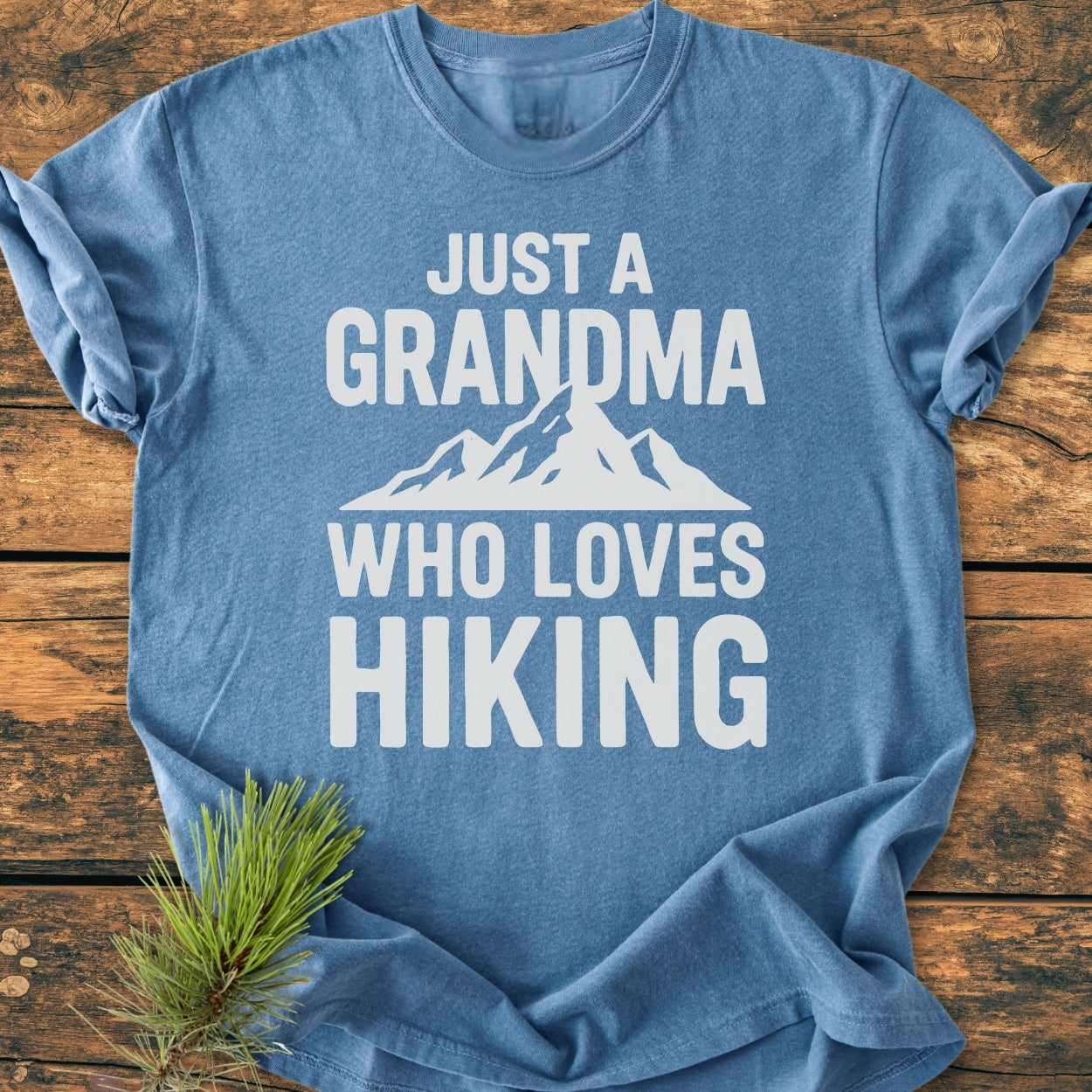Grandma Loves Hiking - Vintage Wash Tee in heather-blue, featuring bold white lettering and a mountain graphic, laid flat on rustic wood with a pine sprig beside it.