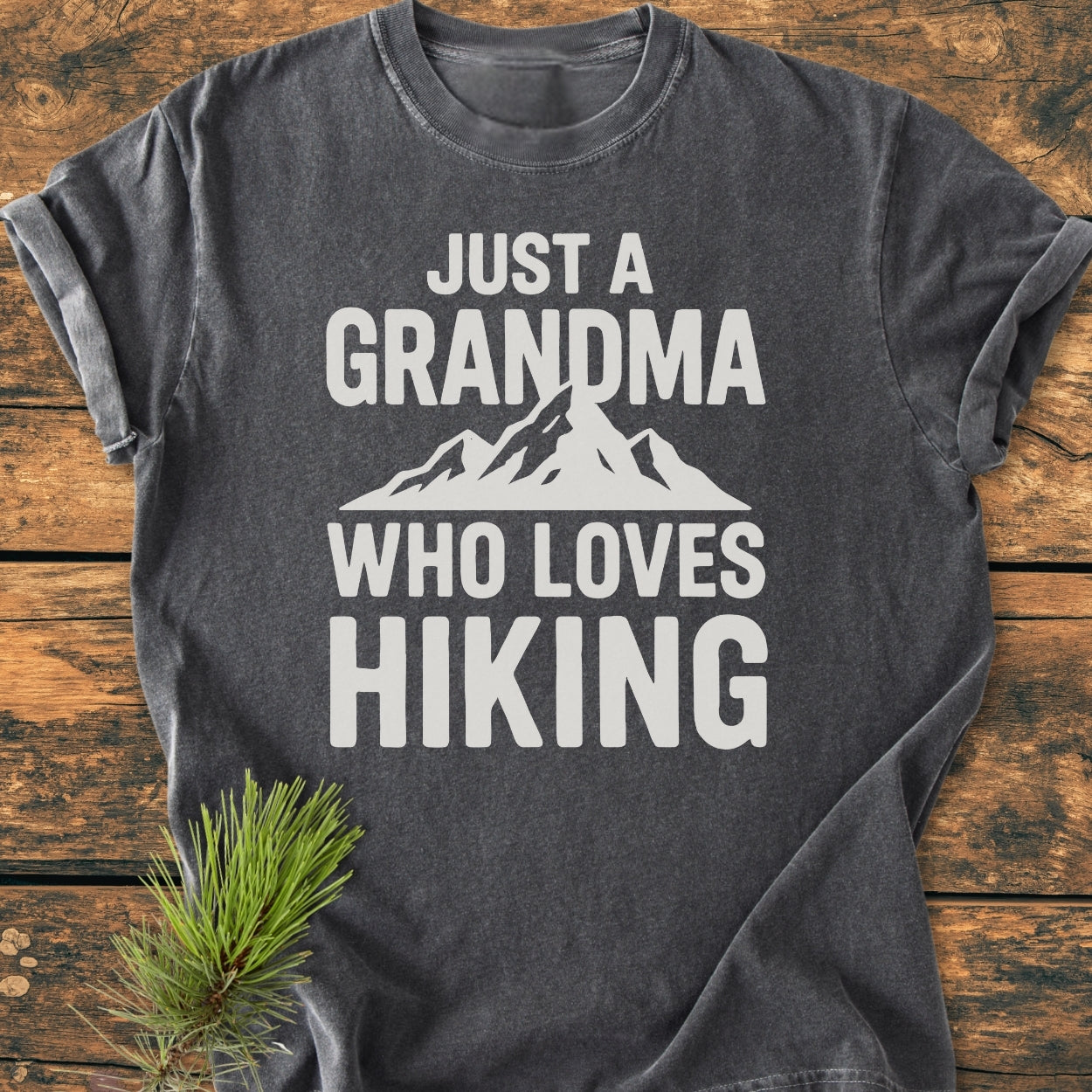Grandma Loves Hiking - Vintage Wash Tee: A dark-gray, short-sleeved T-shirt with the slogan “JUST A GRANDMA WHO LOVES HIKING” and mountain graphic, displayed with a pine sprig on wood.