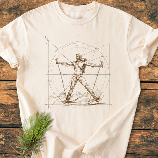 Vitruvian Hiker - Vintage Wash Tee featuring da Vinci-inspired hiker illustration with trekking gear, laid on wood. Includes durable double-needle stitching and soft-washed fabric for comfort. Pine sprig accent nearby.