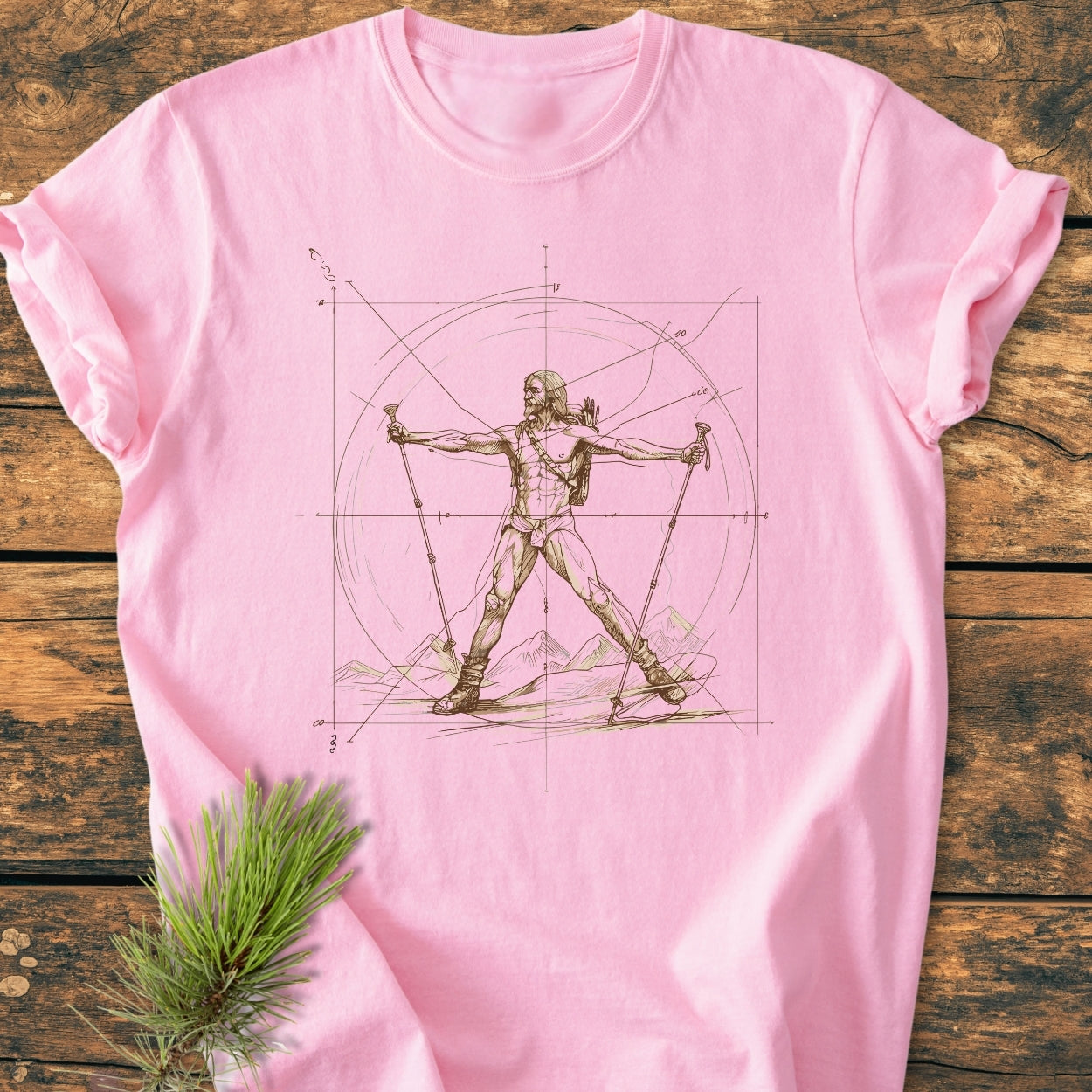 Vitruvian Hiker - Vintage Wash Tee features a sepia-tone Da Vinci-inspired hiker sketch on a soft-pink T-shirt, laid on a wooden surface with a pine sprig, showcasing its relaxed fit and durability.