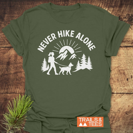 Never Hike Alone Dog Mom T-Shirt featuring a hiking graphic with a dog in a forest, mountains, and sun. Includes NEVER HIKE ALONE text. Soft, durable, pre-shrunk cotton for outdoor enthusiasts.