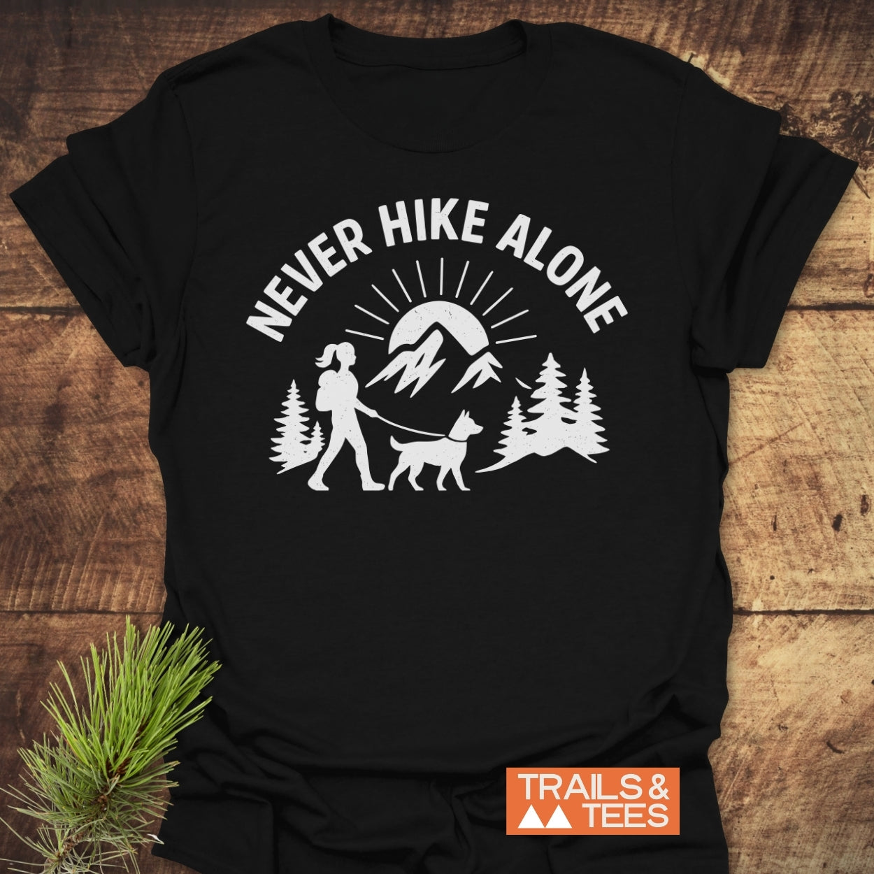 Never Hike Alone Dog Mom T-Shirt featuring a white outdoor scene with a mountain, trees, and a woman with a dog. Crafted from soft, lightweight cotton for comfort and durability.