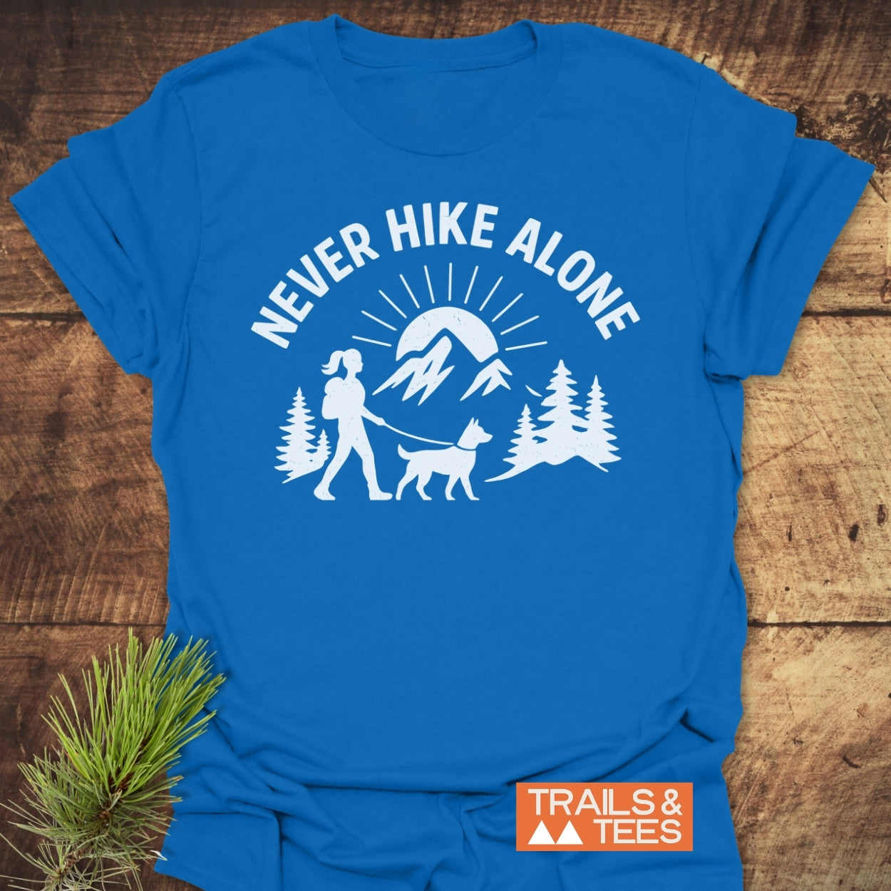 Never Hike Alone Dog Mom T-Shirt featuring a hiker and dog illustration, pine trees, and a rising sun, with the Trails & Tees logo, offering soft, durable comfort for outdoor enthusiasts.