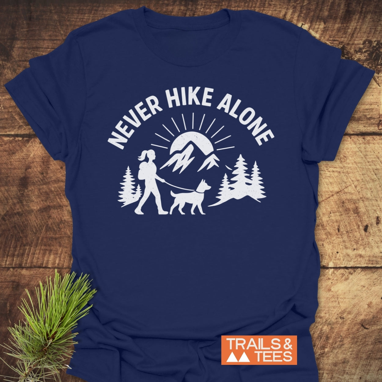 Never Hike Alone Dog Mom T-Shirt featuring a white slogan and graphic of a hiker with dog on navy-blue. Includes TRAILS & TEES label; evergreen sprigs beside for an outdoor theme.