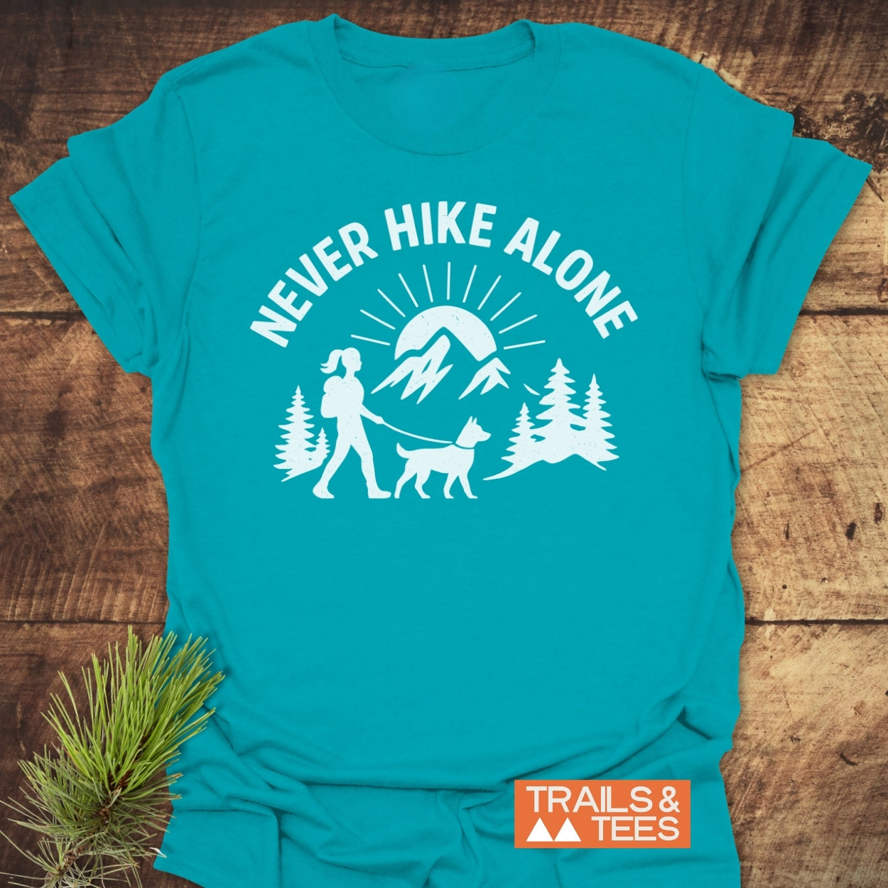 Never Hike Alone Dog Mom T-Shirt features a graphic of a hiker and dog with pine trees and mountains, conveying outdoor adventure. Includes TRAILS & TEES logo, highlighting its trail-tested design.