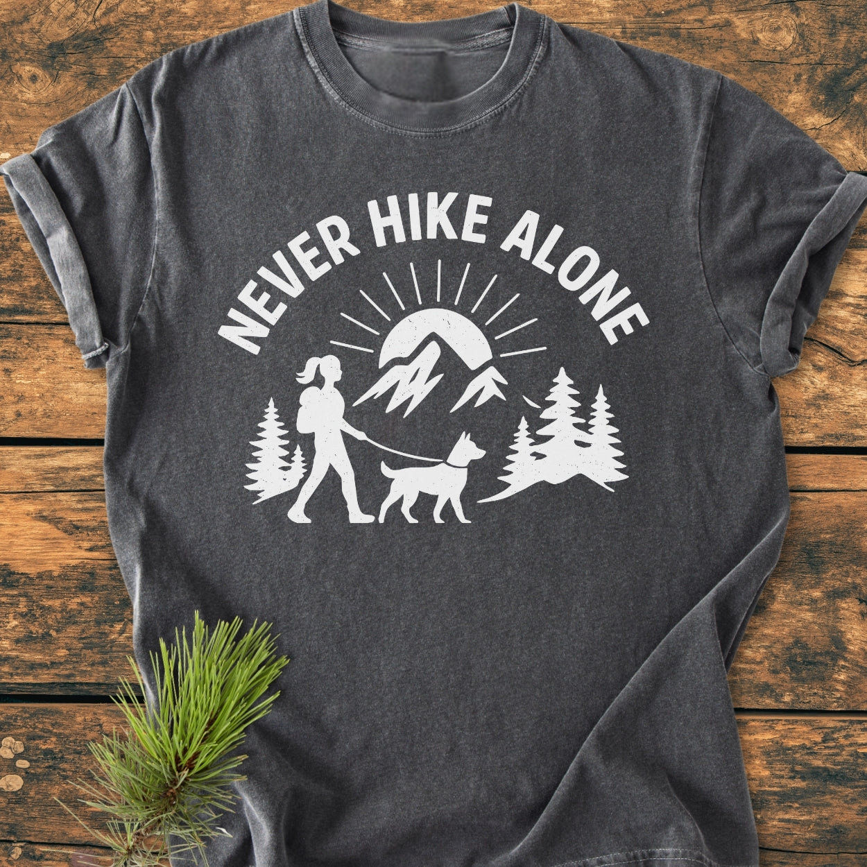 Never Hike Alone Dog Mom - Vintage Wash Tee features a hiker and dog graphic amidst nature, on a charcoal-gray T-shirt, laid flat with pine decor, highlighting its cozy, durable design.