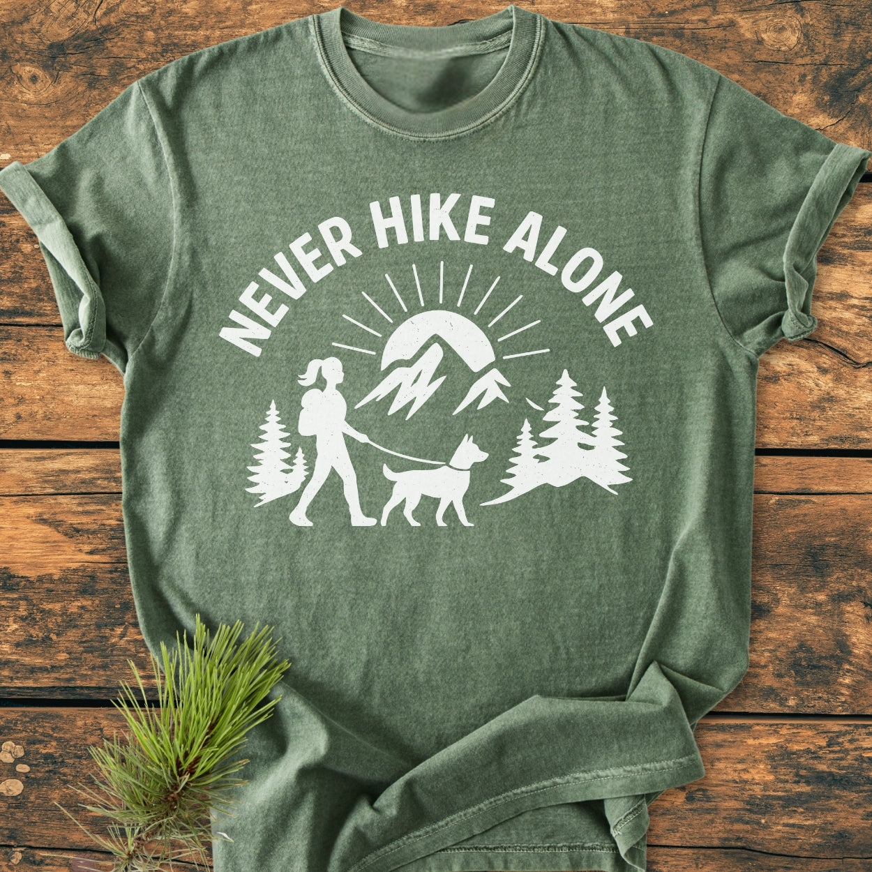 Never Hike Alone Dog Mom - Vintage Wash Tee, in a heather-green hue, showcases a slogan and outdoor scene print, laid on rustic wood with pine needles, highlighting its cozy, relaxed fit.