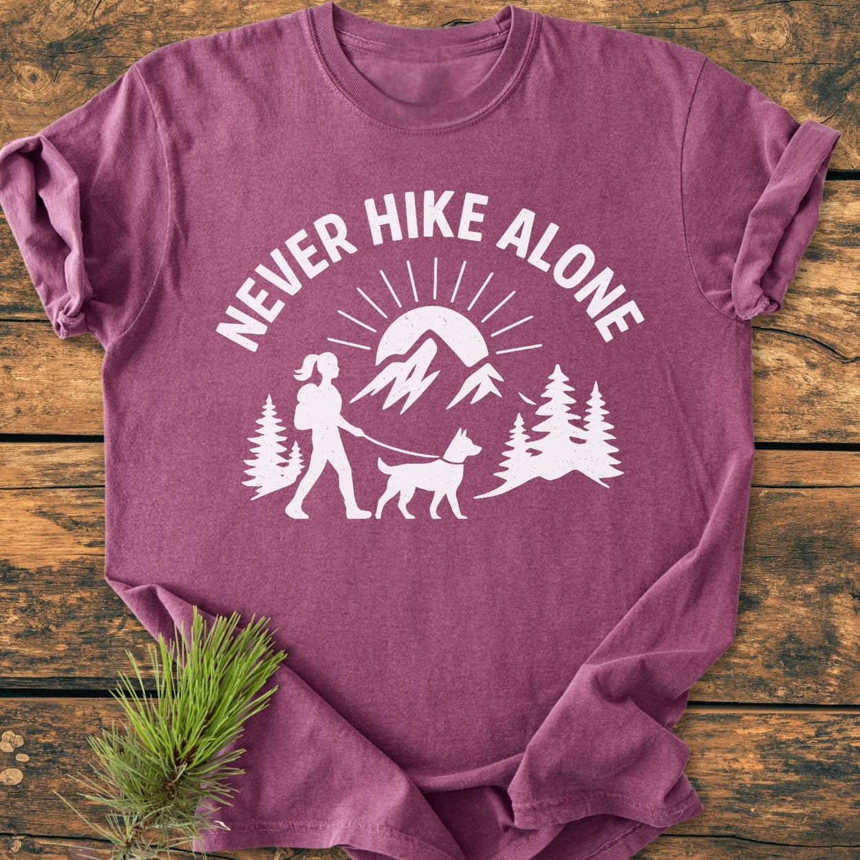 Never Hike Alone Dog Mom - Vintage Wash Tee, featuring a stylized outdoor scene with a hiker, dog, pine trees, and mountains, printed on a heathered purple/mauve crew-neck t-shirt.