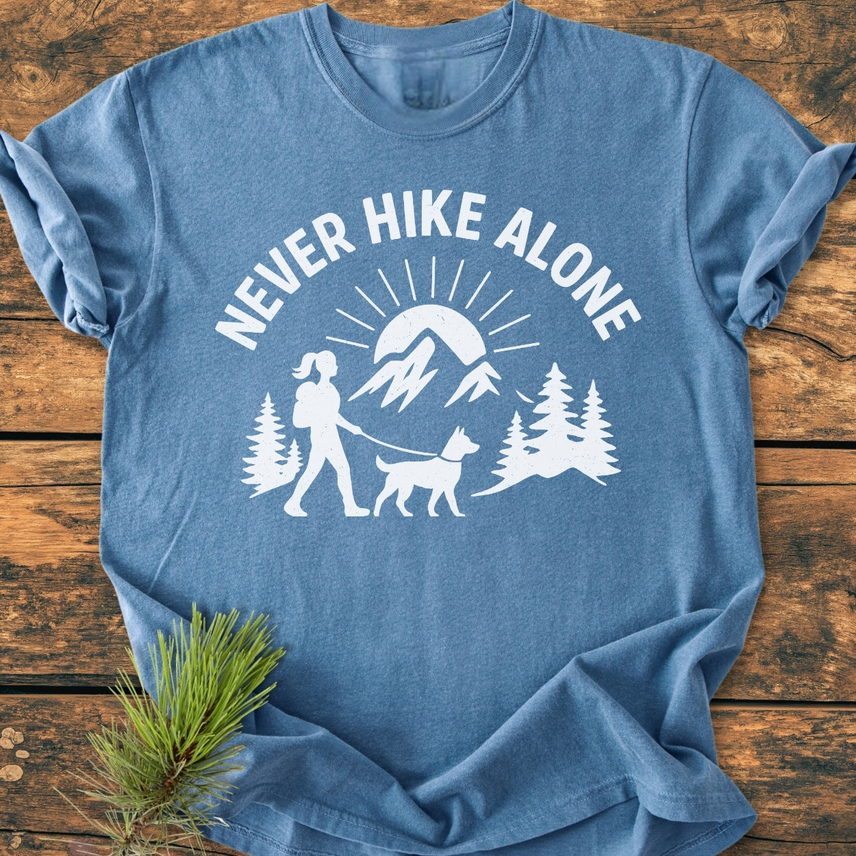 Never Hike Alone Dog Mom - Vintage Wash Tee features a stylized scene with a hiker and dog among trees, mountains, and sun. Made of 100% ring-spun cotton, offering a relaxed fit.