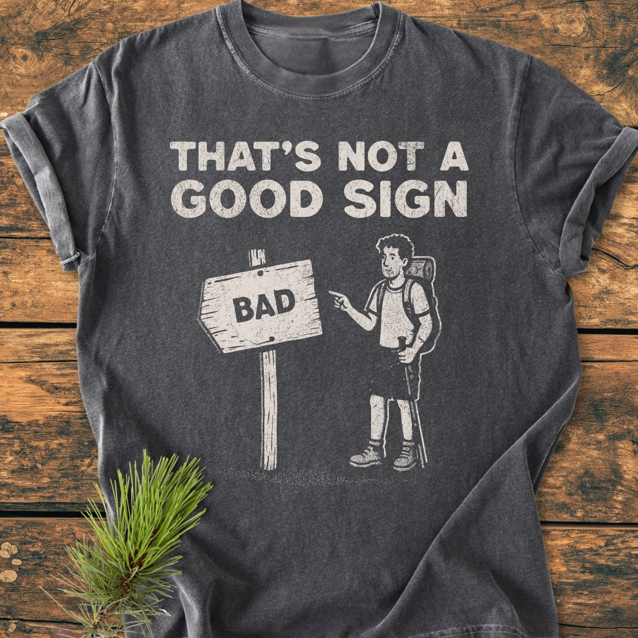 Bad Sign - Vintage Wash Tee: A dark-gray, washed-out T-shirt, featuring a humorous graphic of a backpacker and signpost. Laid on wood, with a pine sprig accentuating its rustic appeal.