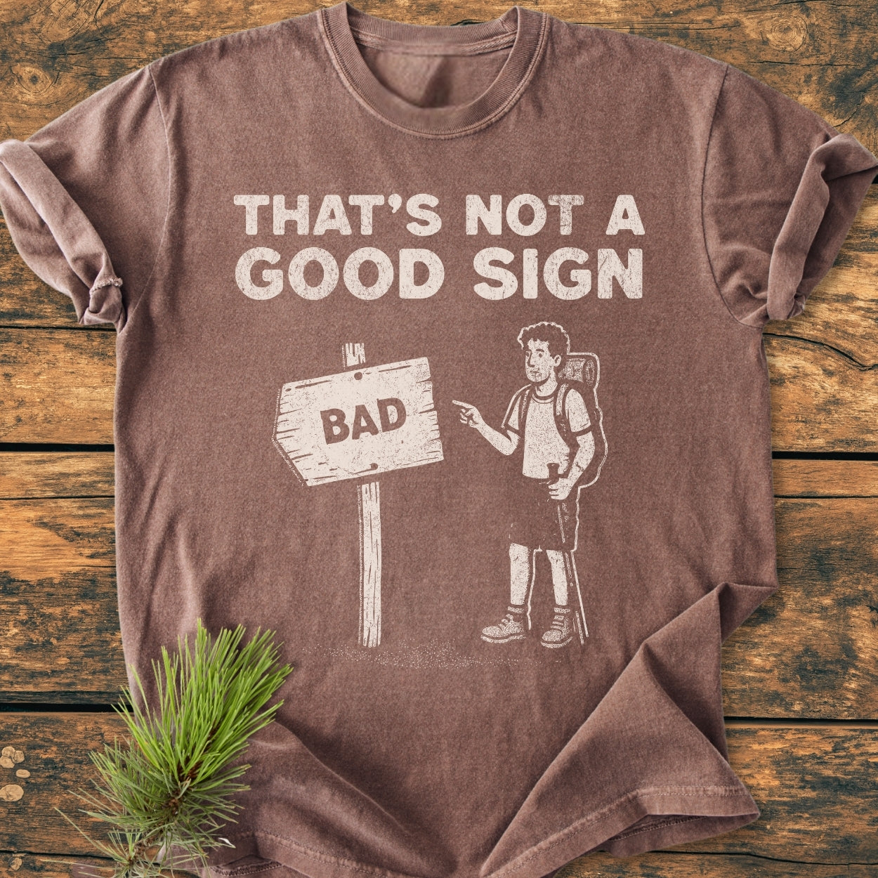 Bad Sign - Vintage Wash Tee featuring a cartoon hiker and witty THAT'S NOT A GOOD SIGN text, displayed flat on a wooden surface. Made from soft, durable 100% ring-spun cotton.