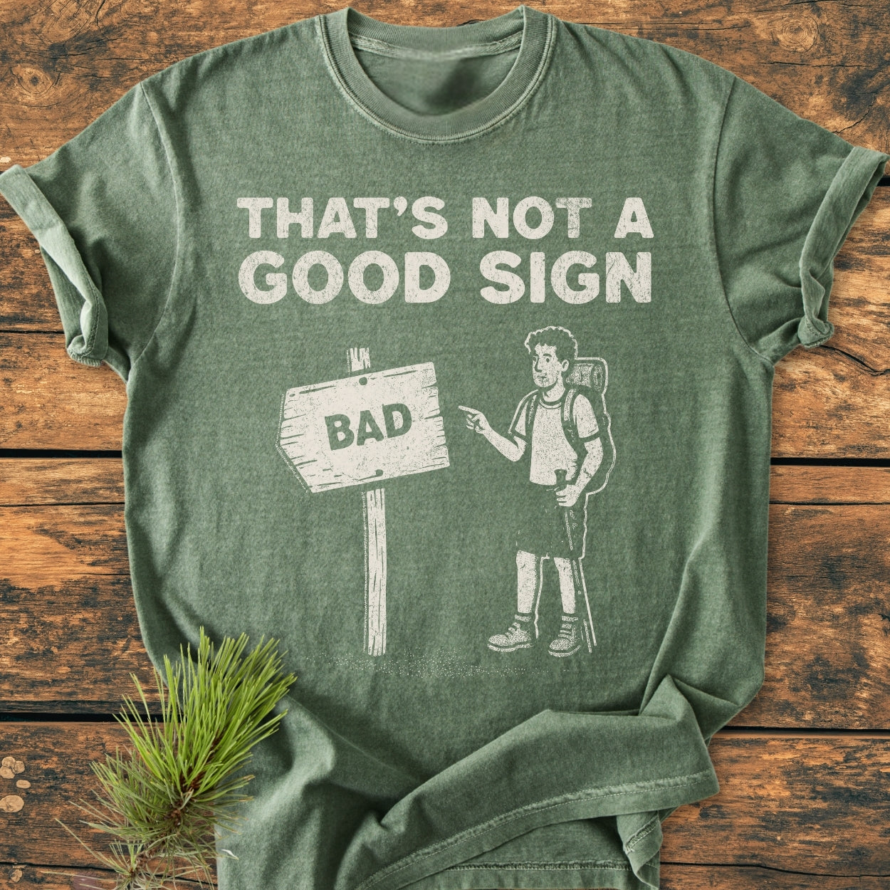 Bad Sign - Vintage Wash Tee features a cartoon hiker with trekking poles pointing at a “BAD” sign. The bold text above reads, “THAT’S NOT A GOOD SIGN.” Perfect for hiking enthusiasts.