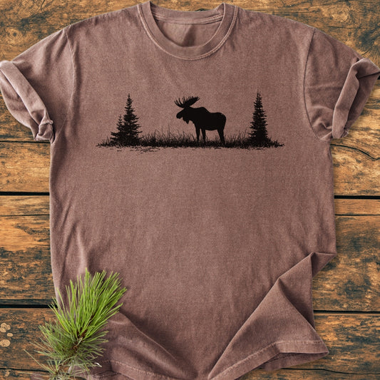 Lone Moose - Vintage Wash Tee laid flat, featuring a black moose and pine tree graphic on stone-washed brown fabric, with a pine sprig accent on a wooden surface.