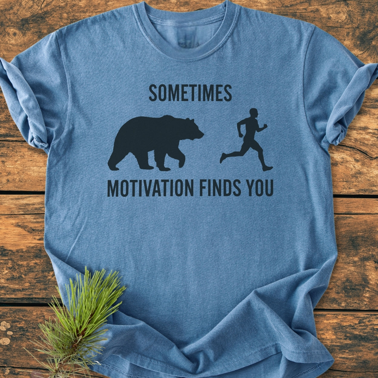 The Sometimes Motivation Finds You - Vintage Wash Tee features a heather-blue color and playful bear-person silhouette design, showcasing a relaxed fit on a wooden backdrop with a pine sprig accent.