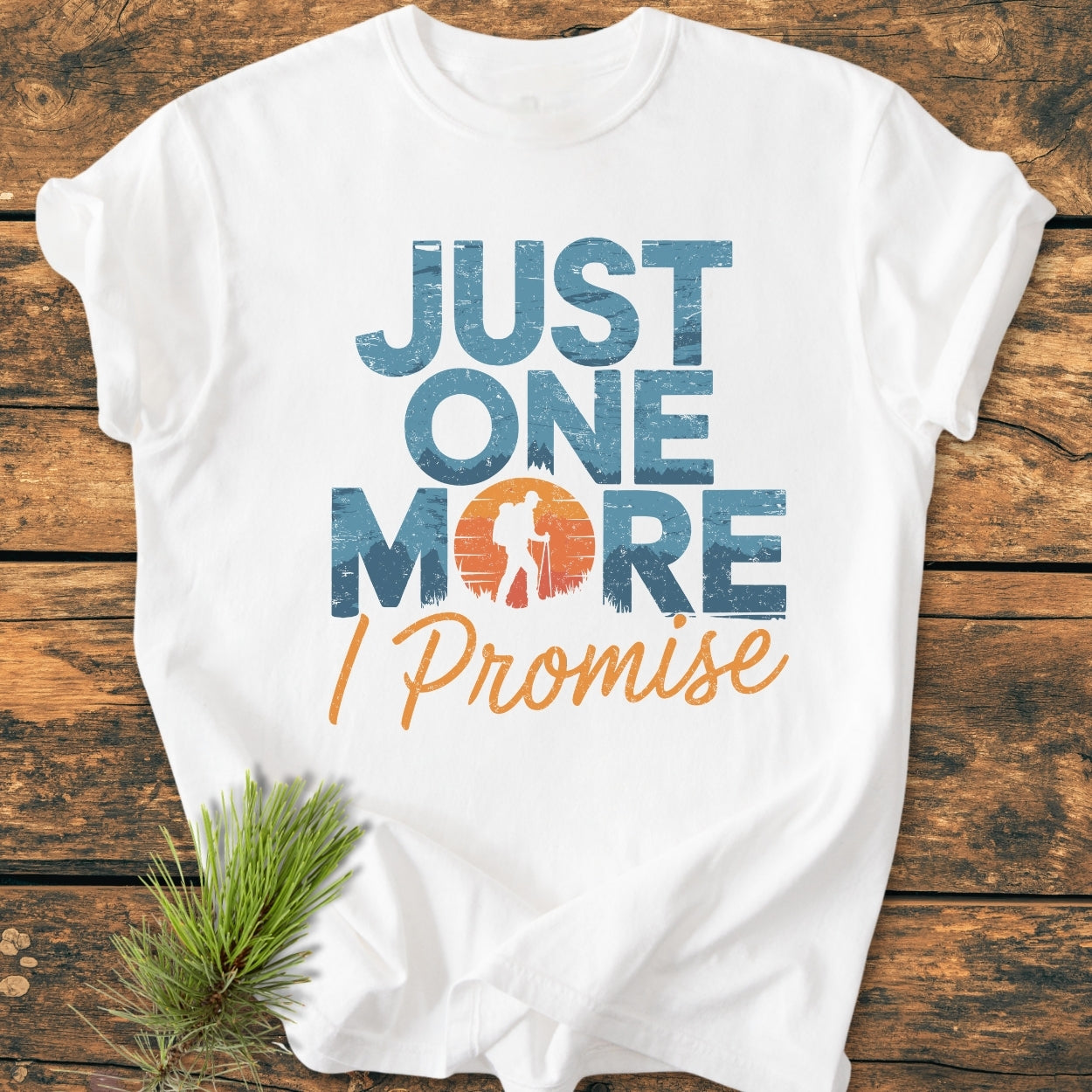 Just One More I Promise - Vintage Wash Tee featuring distressed text and hiker silhouette sunset graphic on a soft, relaxed fit, white garment-dyed tee, laid on wood with pine sprig accent.
