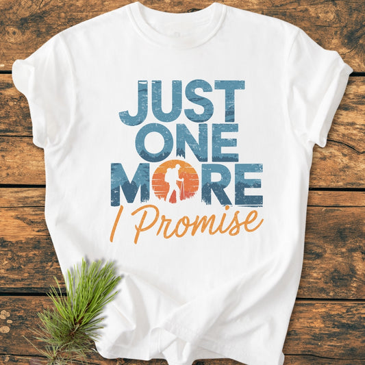 Just One More I Promise - Vintage Wash Tee featuring distressed text and hiker silhouette sunset graphic on a soft, relaxed fit, white garment-dyed tee, laid on wood with pine sprig accent.