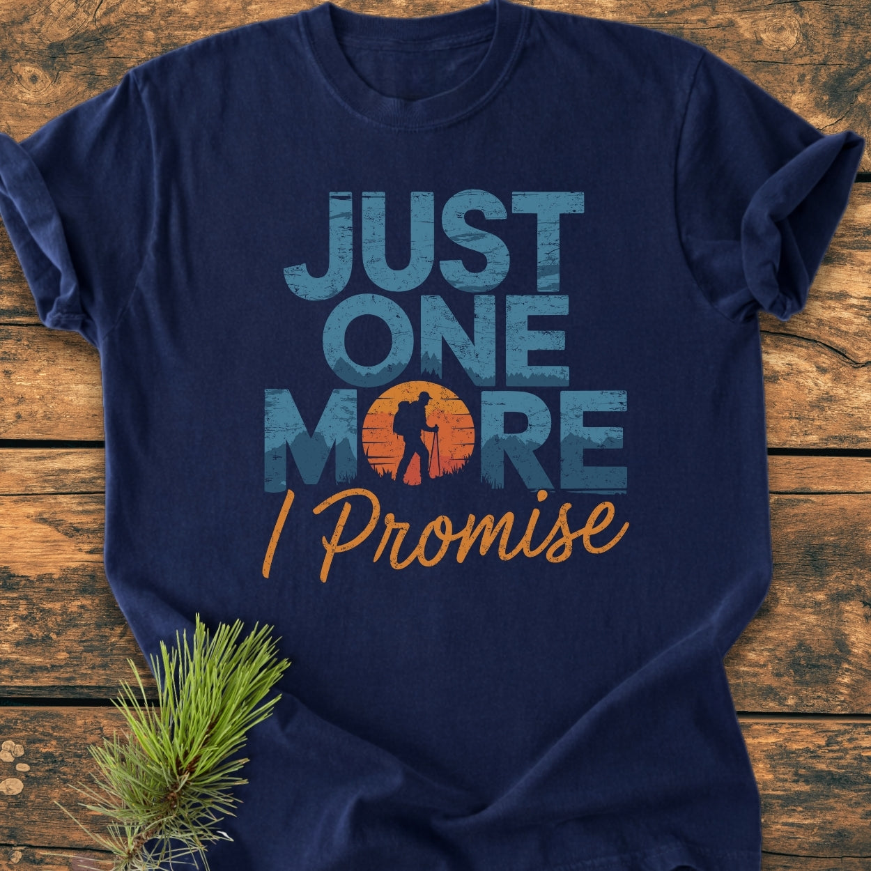 Just One More I Promise - Vintage Wash Tee displayed flat with a hiking-themed graphic and slogan, on a wooden surface, enhanced by a pine sprig to emphasize its outdoorsy appeal.