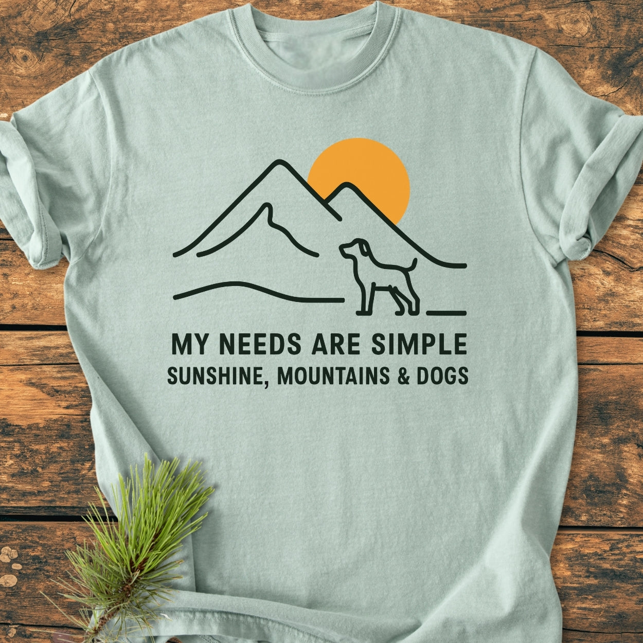 Simple Joys Mountains - Vintage Wash Tee featuring a line-drawing of mountains, sun, and dog, with the slogan MY NEEDS ARE SIMPLE – SUNSHINE, MOUNTAINS & DOGS on a wooden surface with pine sprig.