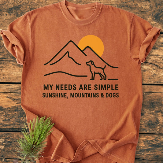 Simple Joys Mountains - Vintage Wash Tee features a black line-drawing of mountains, sun, and dog with the text My needs are simple: Sunshine, mountains & dogs on a rust-orange background.