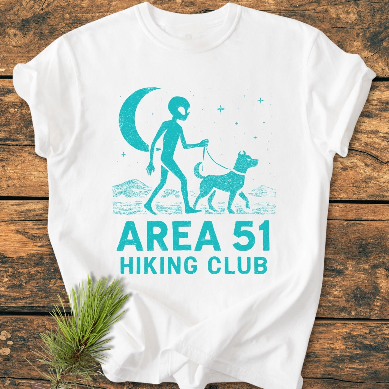 Area 51 Hiking Club - Vintage Wash Tee features a stylized alien walking a dog under a crescent moon. Laid on wood with a pine sprig, showcasing its relaxed fit and soft-washed fabric.