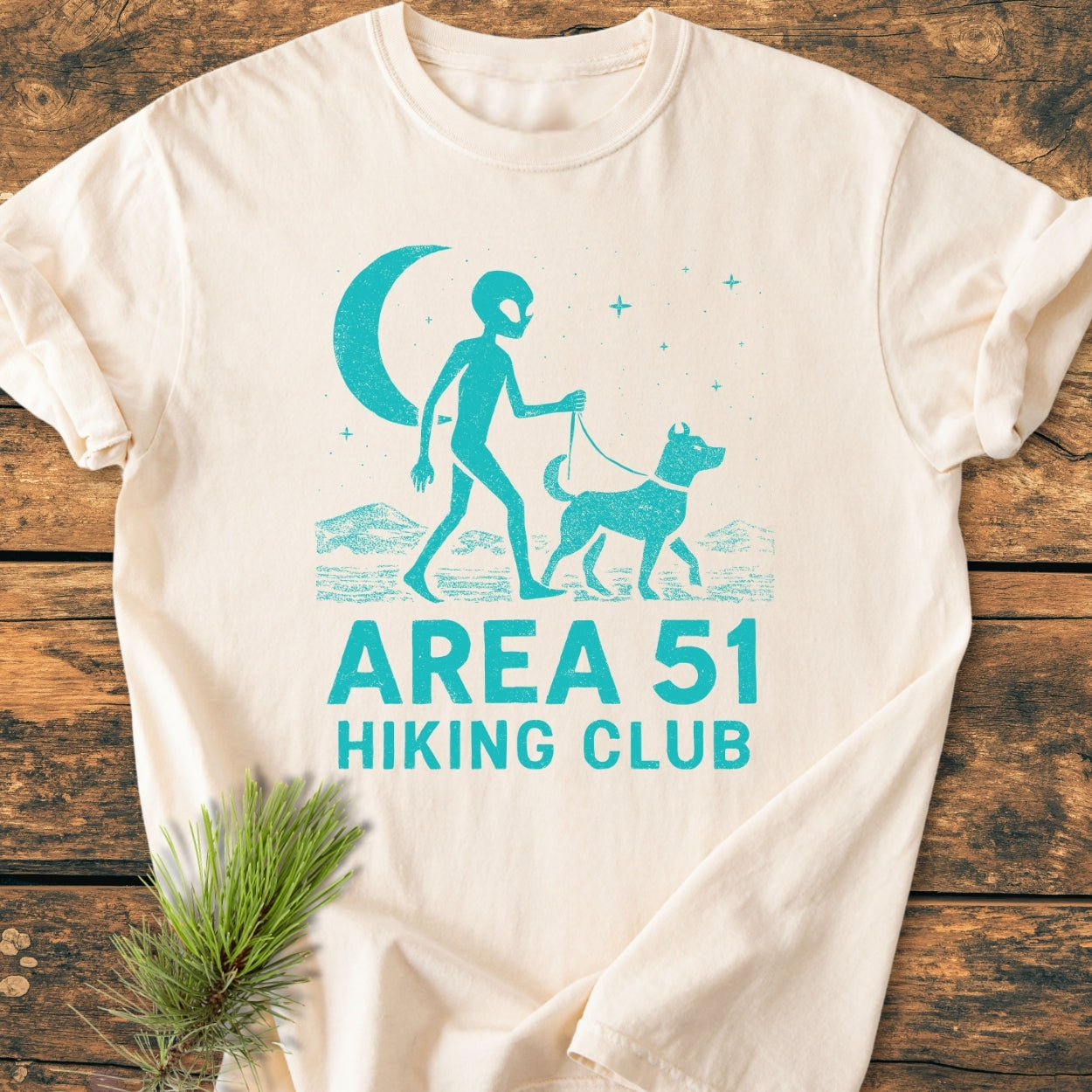 Area 51 Hiking Club - Vintage Wash Tee features a turquoise alien walking a dog under stars and a crescent moon, printed on a light-colored shirt, with a pine sprig accent.