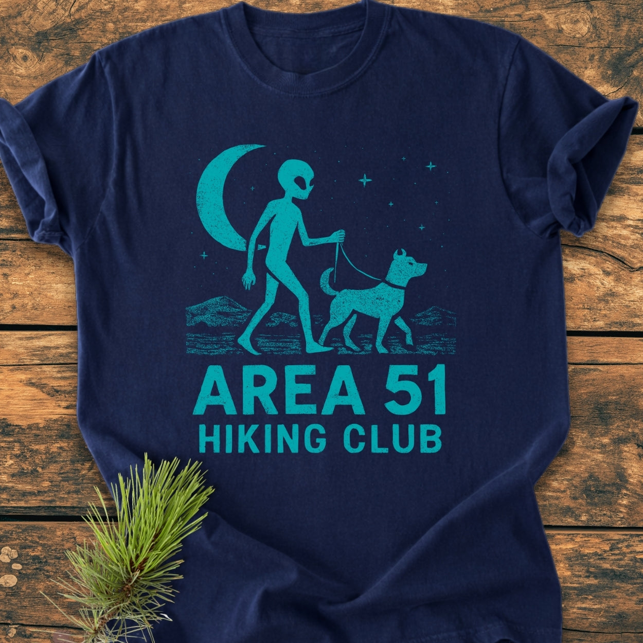 Area 51 Hiking Club - Vintage Wash Tee featuring an alien walking a dog under stars and a crescent moon, printed on a navy-blue shirt, laid on a wooden surface.