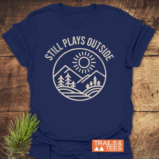 Still Plays Outside T-Shirt on wood with a white mountain graphic, pine trees, and a sun design, styled with a pine sprig; features durable prints and a comfortable fit.
