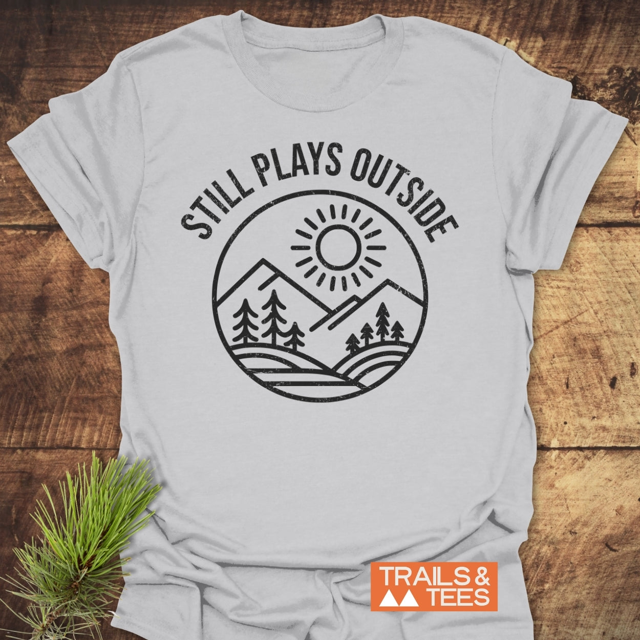 Still Plays Outside T-Shirt with a circular mountain and pine tree design, laid on a wooden surface. Features a small pine sprig and Trails & Tees logo, emphasizing outdoor adventure spirit.