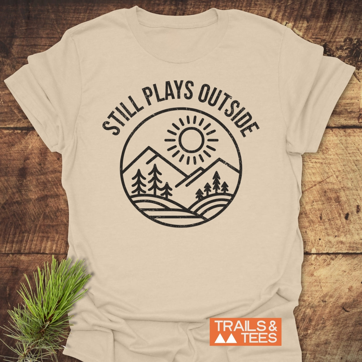 Still Plays Outside T-Shirt featuring a circular design of hills, trees, and sun, laid on wood. Includes Trails & Tees logo and pine sprig, emphasizing outdoor adventure and comfort.