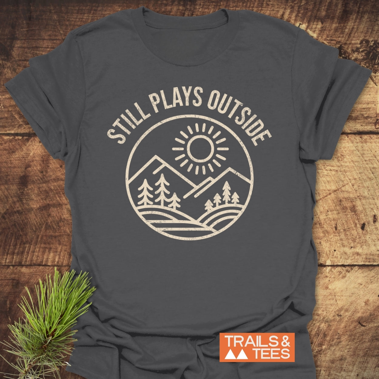 Still Plays Outside T-Shirt featuring a mountain-and-forest graphic on charcoal-gray fabric, laid flat with a pine sprig, exemplifies outdoor adventure and comfort through its soft, durable cotton design.