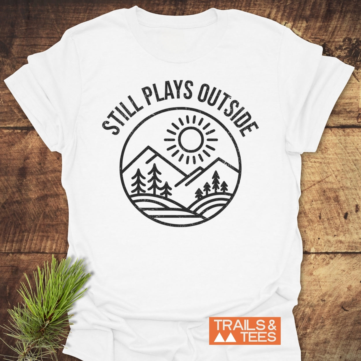 Still Plays Outside T-Shirt featuring a black line-art design of nature on a white tee. Includes TRAILS & TEES logo. Laid on wood with pine needles beside it.