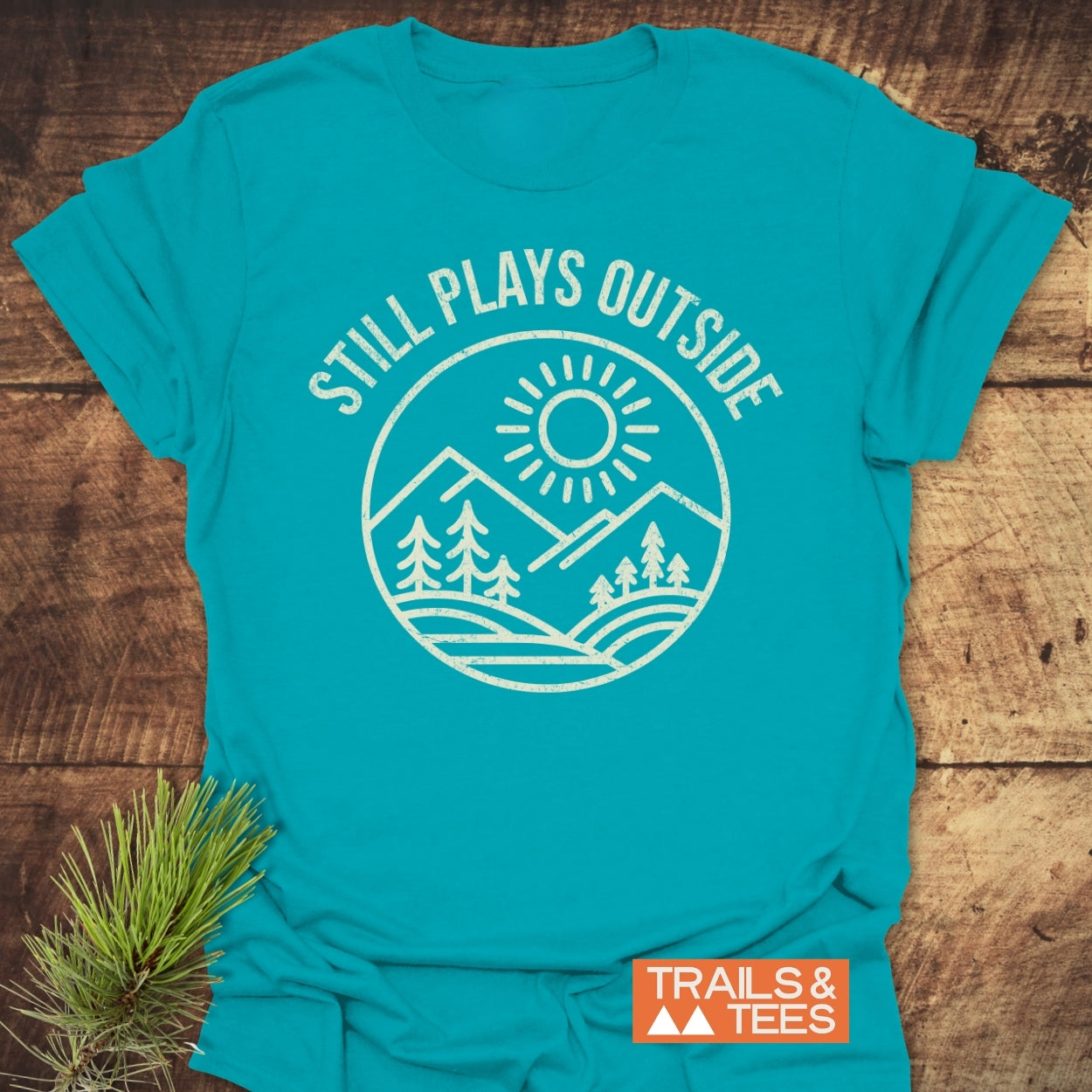 Still Plays Outside T-Shirt featuring a white sun and mountain graphic, laid flat on a wooden surface. Crafted from soft cotton, ideal for outdoor enthusiasts, emphasizing comfort and durability.