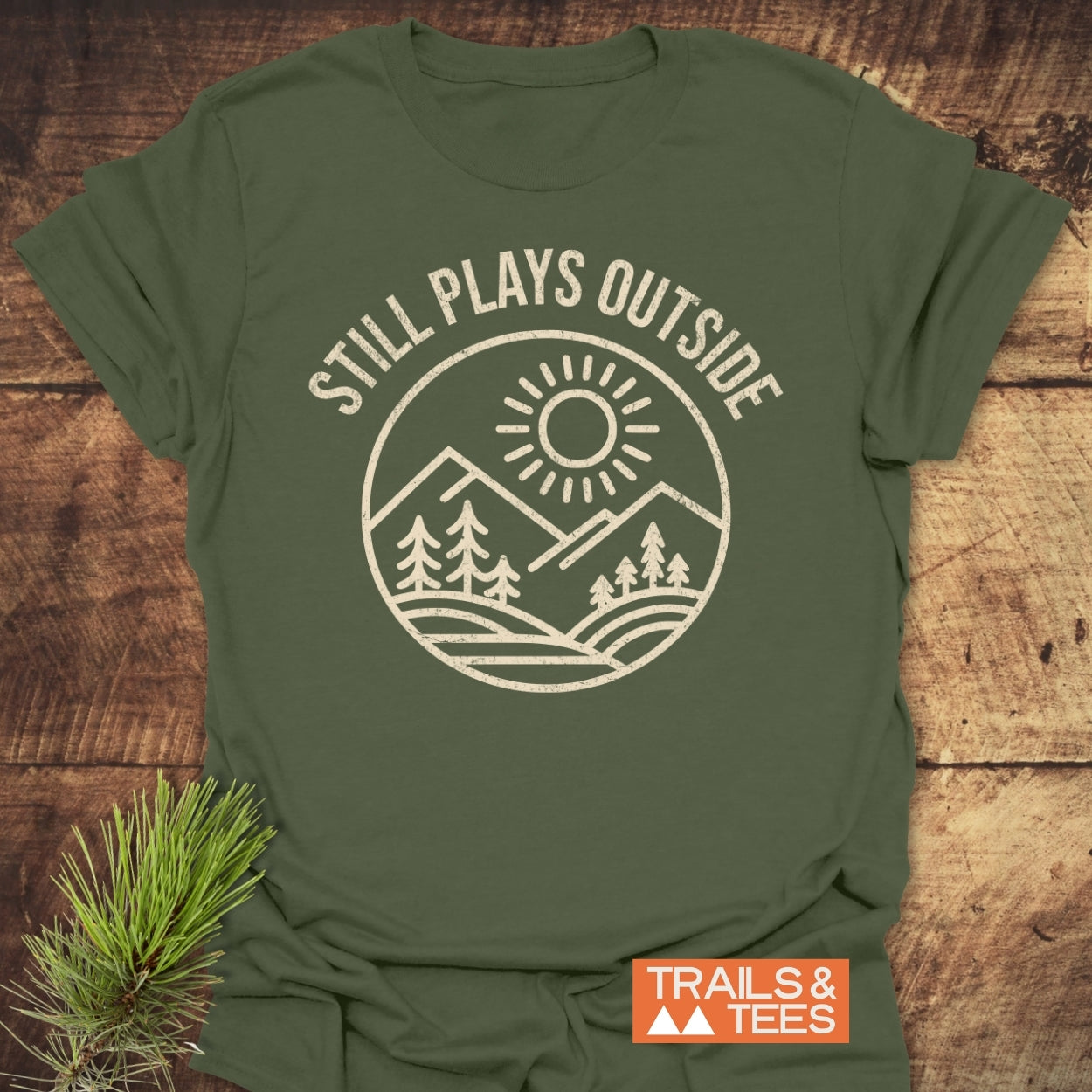 Still Plays Outside T-Shirt with a white line drawing of nature on olive-green fabric. Features STILL PLAYS OUTSIDE text, orange TRAILS & TEES logo, and pine sprig detail.