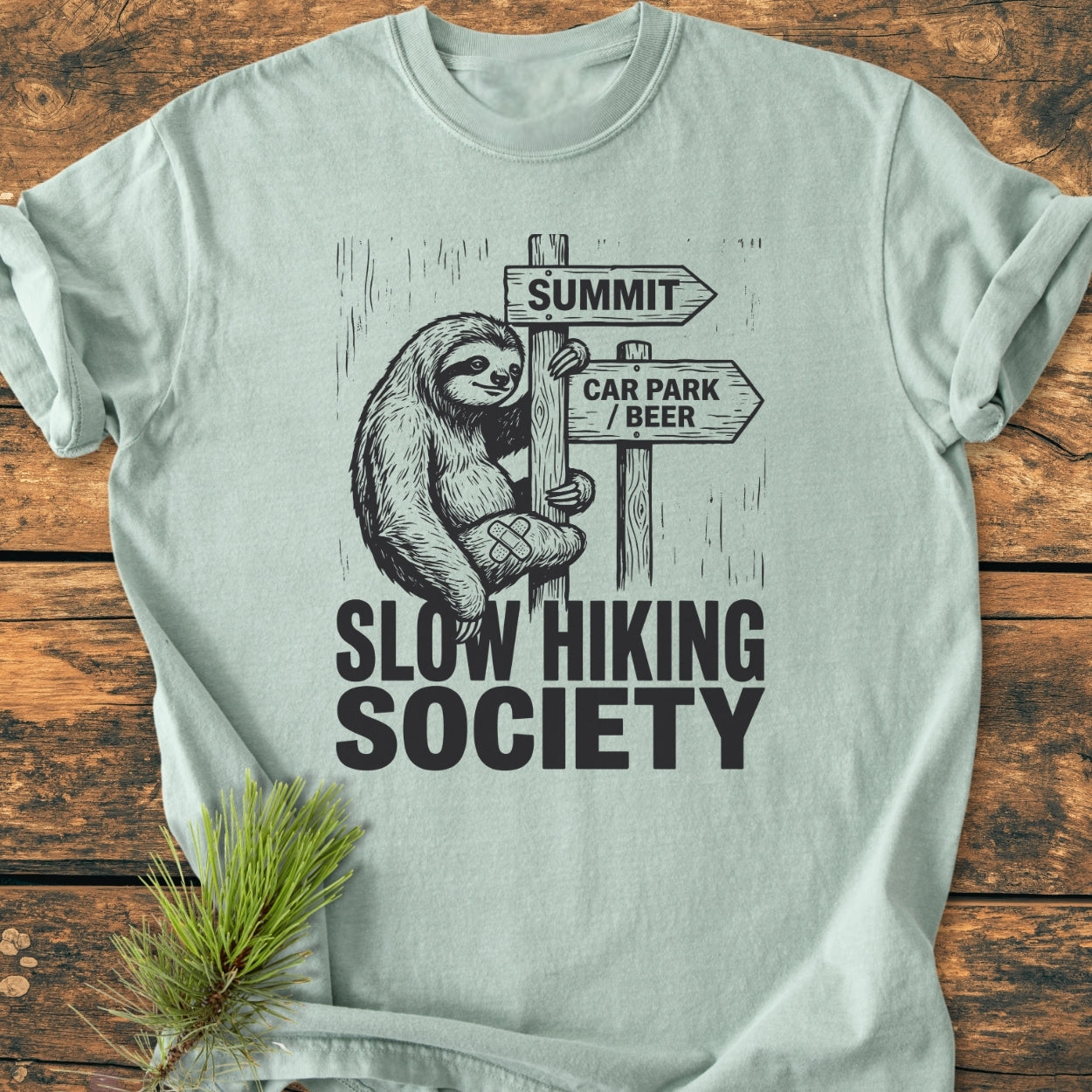 Slow Hiking Society - Vintage Wash Tee features a cartoon sloth on a signpost saying SUMMIT and CAR PARK / BEER, laid on a wooden surface with a pine sprig accent.