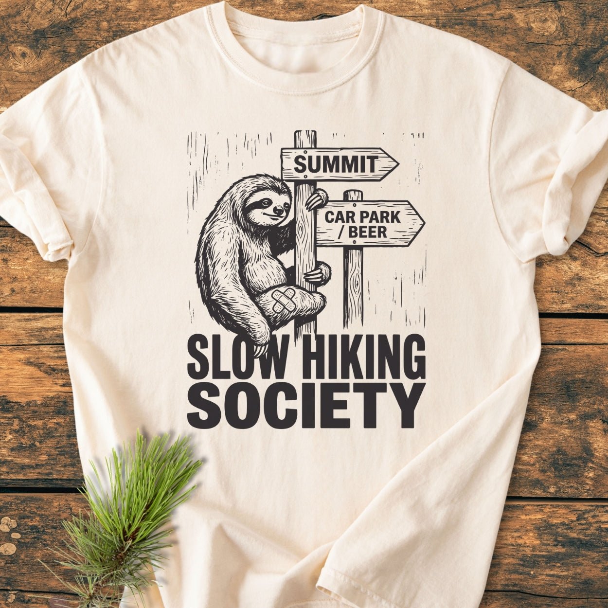 Slow Hiking Society - Vintage Wash Tee featuring a smiling sloth on a signpost with arrows. Soft, garment-dyed fabric offers a relaxed fit, laid on a wooden surface with a pine sprig.