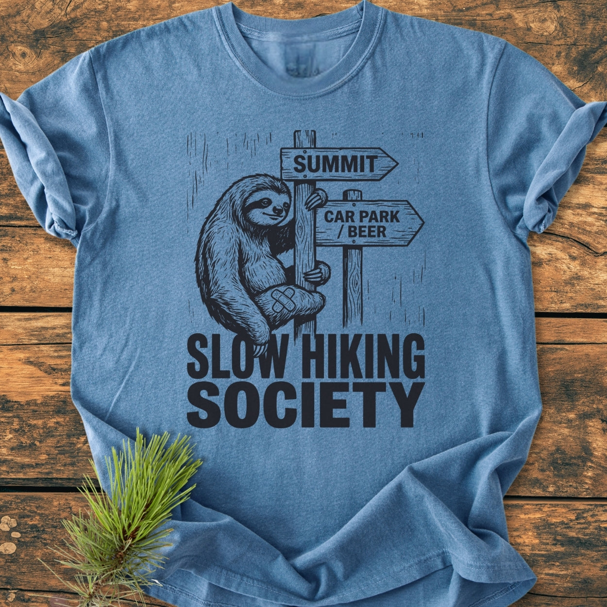 Slow Hiking Society - Vintage Wash Tee featuring a cartoon sloth on a signpost, laid on wood with pine sprig, showcases relaxed fit and durable stitching, emphasizing comfort and style.