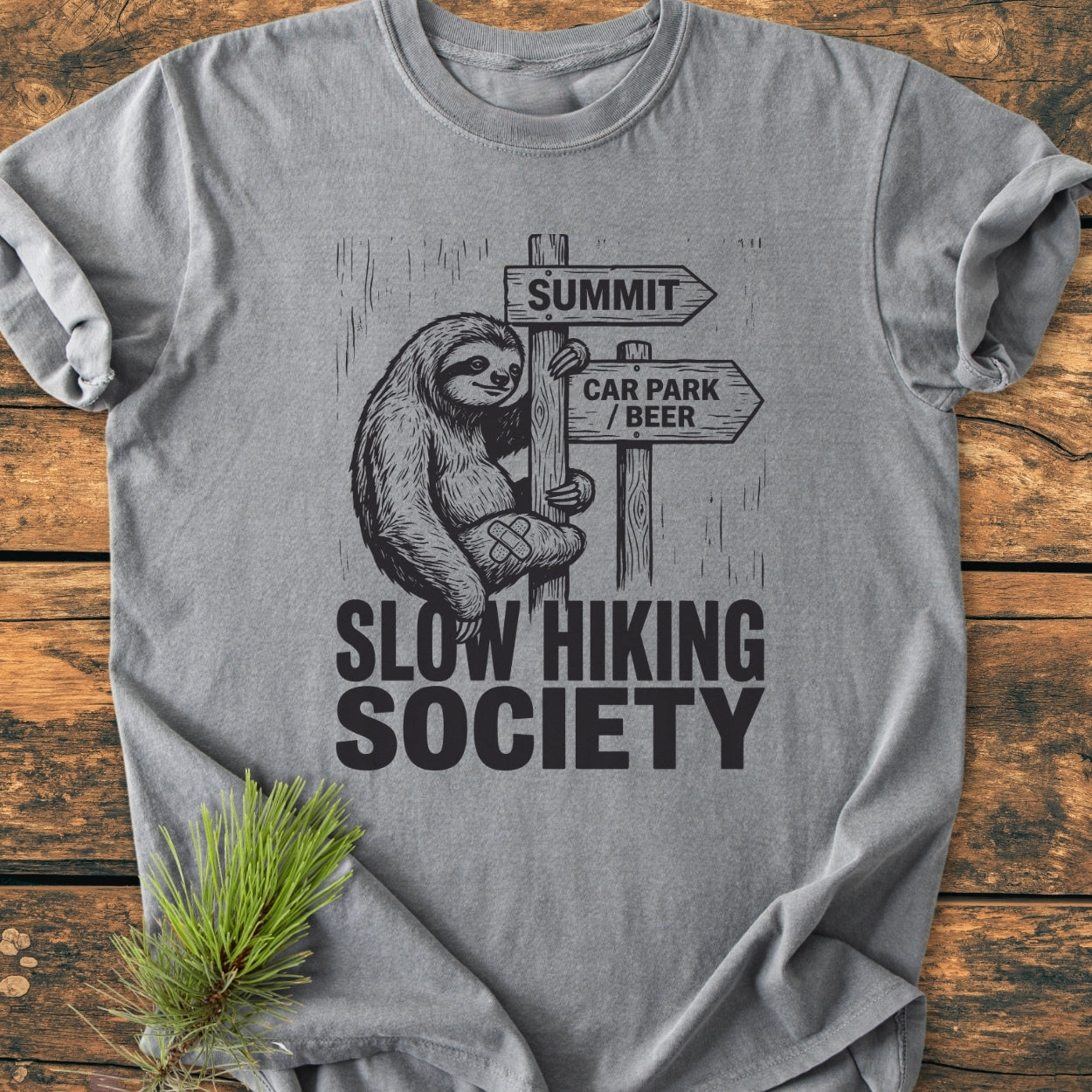 Slow Hiking Society - Vintage Wash Tee featuring a cartoon sloth on a trail sign, laid on wood, with SUMMIT and CAR PARK / BEER arrows. Accentuated with a pine sprig for styling.