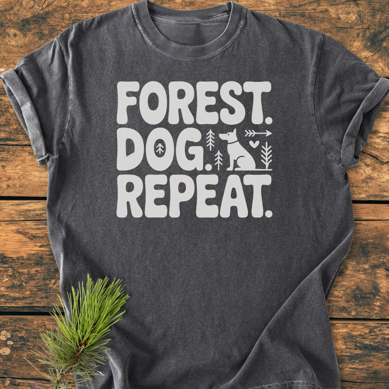 Forest Dog Repeat - Vintage Wash Tee on rustic wood, featuring white text and graphics of a dog and trees. Soft, relaxed fit, showcasing outdoor theme with pine sprig accent.
