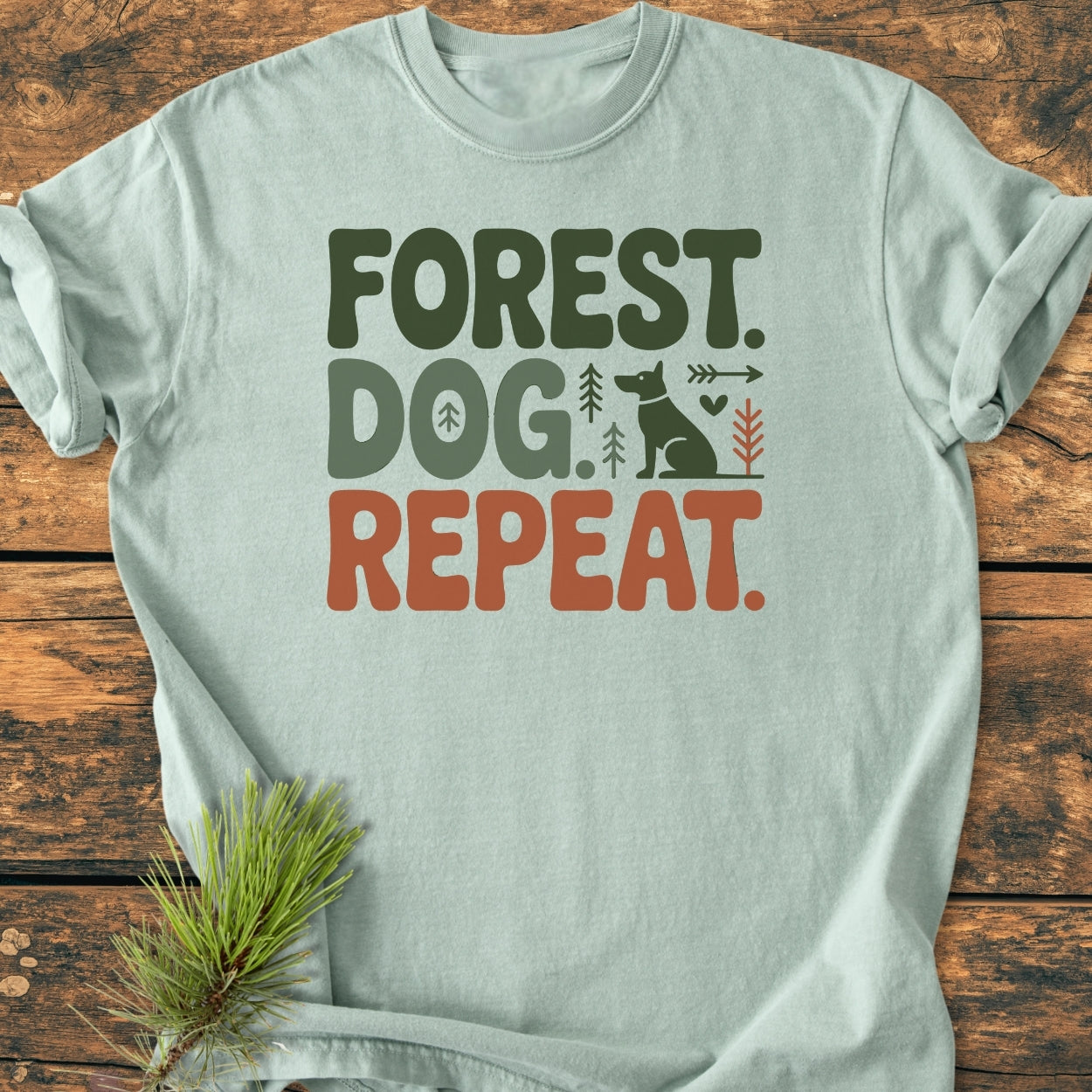 Forest Dog Repeat - Vintage Wash Tee in light-sage green with forest-green and rust-orange text, featuring a dog silhouette, pine trees, arrow, and heart graphic on a wooden surface.