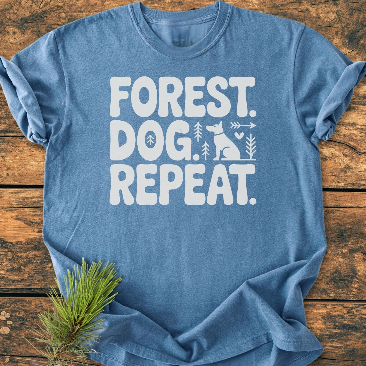Forest Dog Repeat - Vintage Wash Tee on rustic wood, showcasing white FOREST. DOG. REPEAT. text with pine trees, arrow, heart, and dog silhouette graphics; complemented by a pine branch.