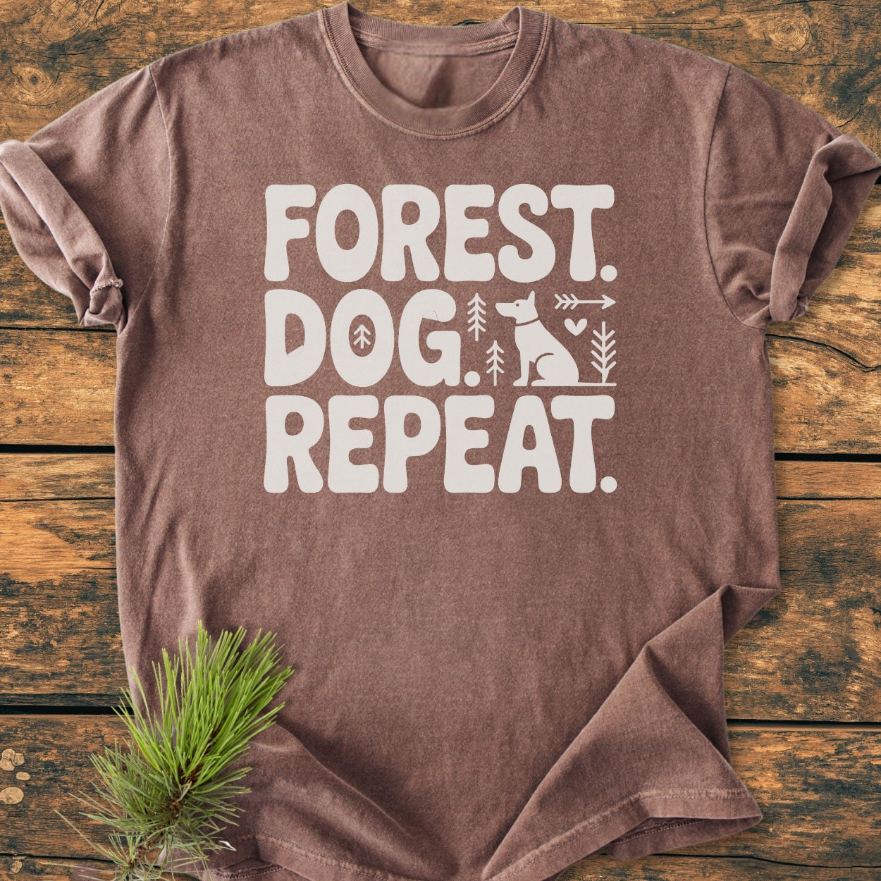 Forest Dog Repeat - Vintage Wash Tee features “FOREST. DOG. REPEAT.” in white with tree, arrow, heart, and dog graphics. Displayed on a wooden surface with pine needles, showcasing its relaxed fit and durable stitching.