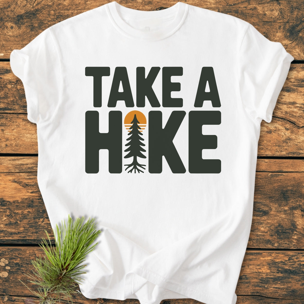 Take A Hike - Vintage Wash Tee features a bold graphic with a tree replacing 'I' in 'HIKE' on a soft, relaxed fit white T-shirt, laid on a wooden surface.