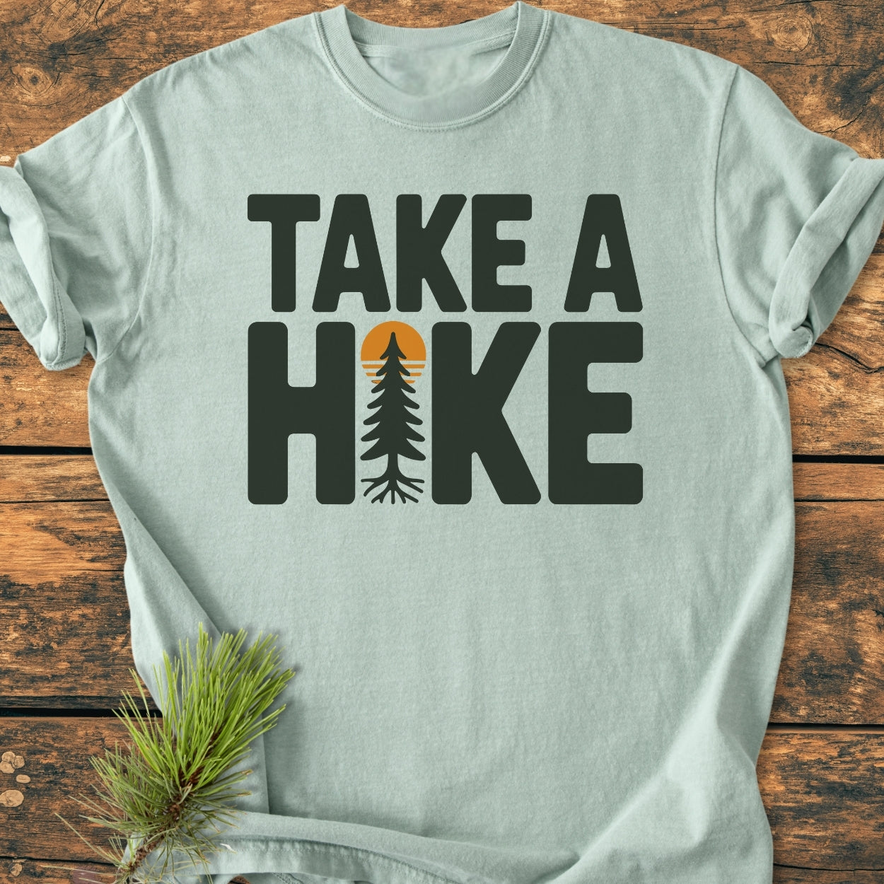 Take A Hike - Vintage Wash Tee features a light-green hue with a bold slogan and stylized fir tree, laid flat on wood, embodying an outdoorsy spirit with pine sprig accent.